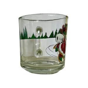 May include: A clear glass mug featuring a festive graphic of a character in a red and white suit skating on ice. The design includes green pine trees in the background and the text Disney printed near the base. The mug has a sturdy handle.