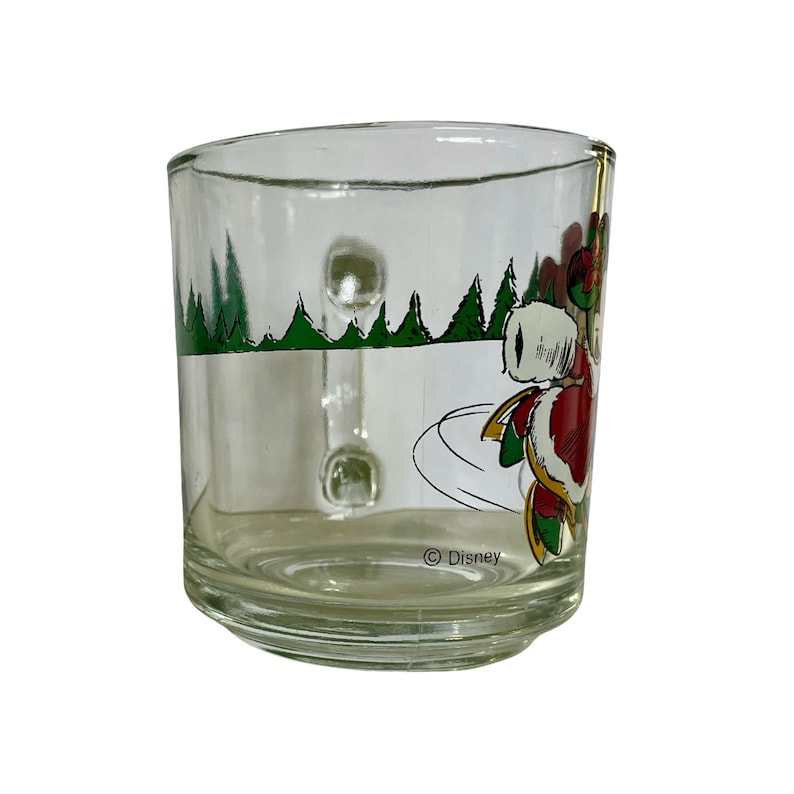 May include: A clear glass mug featuring a festive graphic of a character in a red and white suit skating on ice. The design includes green pine trees in the background and the text Disney printed near the base. The mug has a sturdy handle.