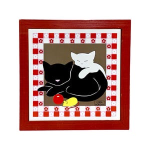 May include: A framed tile with a red checkered border and a brown background. Two cats, one black and one white, are curled up together. The black cat has a red ball and a yellow mouse toy. The tile is signed "Linda Morgan".