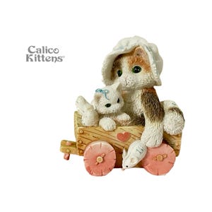 May include: A figurine of two kittens in a wooden wagon with pink wheels. One kitten wears a bonnet and the other has a blue bow. A small mouse is in front of the wagon. The words "Calico Kittens" are on the left.