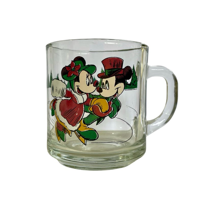 May include: A clear glass mug featuring a colorful graphic of Mickey and Minnie Mouse ice skating. The characters wear festive winter attire including hats and coats, set against a background of green pine trees on a white ice surface.