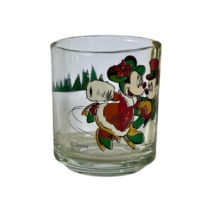May include: A clear glass tumbler featuring a colorful graphic of Mickey and Minnie Mouse ice skating in winter attire. The design includes green pine trees, red and green clothing, and the text Disney printed near the base of the glass.