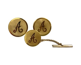 May include: A set of gold-toned cufflinks and tie tack. Each piece features the letter "A" in a script font. The cufflinks are round, while the tie tack has a chain and a bar closure. The set is likely vintage.