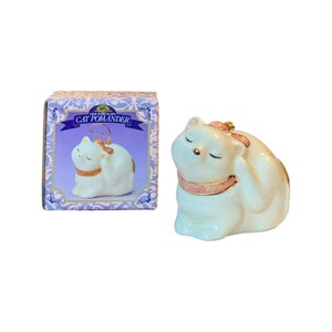 May include: White porcelain cat figurine with pink and gold accents. The cat is curled up with its head resting on its paw. The figurine is a pomander, which is a type of scented ornament. The box reads "Fine Porcelain Cat Pomander".