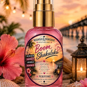 May include: A clear glass spray bottle containing pink liquid with a gold cap. The label reads Mermaid & Mangrove Apothecary, Boom Shakalaka, and lists scent notes of raspberry, strawberry, citrus orange, and tropical, with a net weight of 1 ounce.