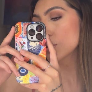 May include: A patterned phone case with a fruit and stamp design. The case features a variety of fruits, stamps, and other decorative elements. The phone case is held in front of a person's face.