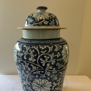 Puede incluir: Un jarrón de cerámica azul y blanco con tapa. El jarrón presenta un patrón floral en tonos azules y blancos. La tapa tiene un pequeño pomo y un borde decorativo. El jarrón probablemente se utiliza para almacenamiento o decoración.