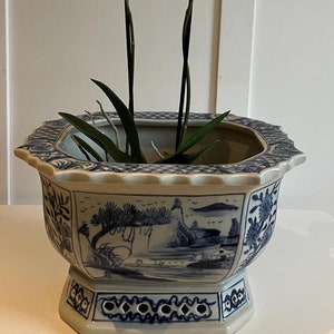 May include: A white ceramic planter with a blue and white floral design. The planter has a square base and a round top. It is decorated with a scene of a mountain range and a pine tree. The planter is filled with a green plant and two white orchids with purple spots.