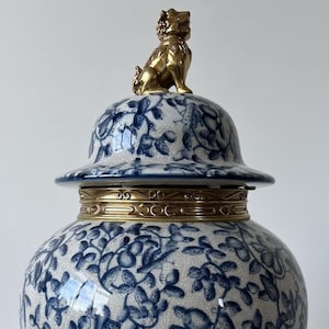 Puede incluir: Un gran jarrón de porcelana azul y blanco con un remate de león dorado y una base dorada. El jarrón está decorado con un patrón floral.
