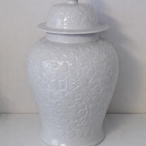 May include: A white ceramic ginger jar with a removable lid featuring a pointed finial. The entire exterior surface of the jar and the lid is decorated with a raised, monochromatic floral pattern that covers the body from the base to the top.