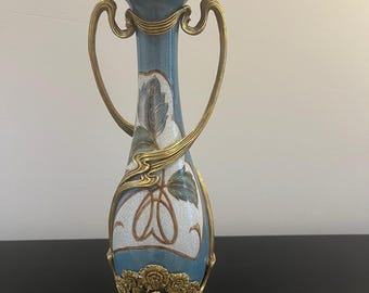 Op de afbeelding: Een hoge, slanke keramische vaas met een lichtblauwe en witte craquelé glazuurafwerking. De vaas is versierd met sierlijke goudkleurige metalen accenten, waaronder twee gebogen handvatten en een decoratieve bloemenvoet met complexe bladmotieven.