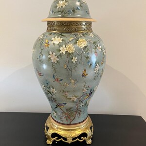 Op de afbeelding: Een grote keramische gemberpot met een lichtblauw-groene basis, voorzien van handgeschilderde witte en gele bloemmotieven, kleurrijke vlinders en vogels. De pot heeft een bijpassend deksel met een goudkleurige knop en een decoratieve metalen voet.
