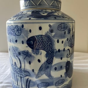 Könnte beinhalten: Ein blau-weißes Keramikgefäß mit Deckel. Das Gefäß ist mit Fischen, Wasserpflanzen und Wellenmustern verziert. Der Deckel hat ein geometrisches Design. Das Gefäß ist zylindrisch und hat eine abgerundete Oberseite.