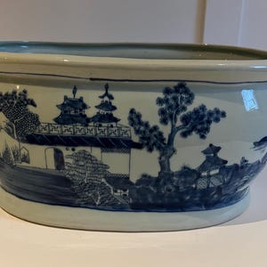 Op de afbeelding: Een grote, ovale keramische plantenbak met een blauw-wit ontwerp. De plantenbak heeft een traditionele Chinese landschapsscène met gebouwen, bomen en figuren.