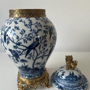 May include: A blue and white porcelain vase with a gold lid and base. The vase is decorated with a floral pattern and birds. The lid features a gold lion finial.