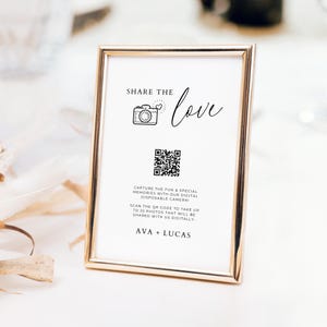 May include: A white sign with a gold frame, with the text "SHARE THE love" and a camera icon. The sign also includes a QR code with the text "CAPTURE THE FUN & SPECIAL MEMORIES WITH OUR DIGITAL DISPOSABLE CAMERA! SCAN THE QR CODE TO TAKE UP TO 25 PHOTOS THAT WILL BE SHARED WITH US DIGITALLY. AVA + LUCAS"