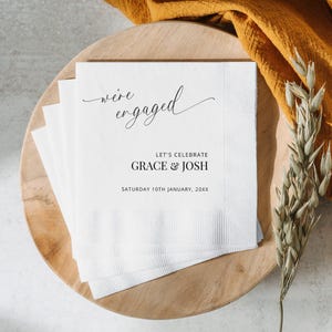 May include: White paper napkins with black text that reads "we're engaged" in cursive, "Let's Celebrate Grace & Josh" and "Saturday 10th January, 20XX".