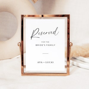 May include: A gold-framed sign with the text "Reserved for the Bride's Family" and the names "Ava + Lucas" in black lettering.
