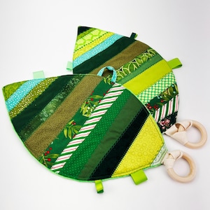 May include: Two leaf-shaped baby sensory toys in shades of green, featuring diverse fabric patterns like stripes and polka dots. Each toy has a wooden ring attached with cream fabric. Green ribbon loops are sewn around the edges.