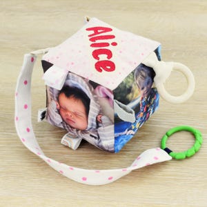 Puede incluir: Un cubo de bebé suave con fotos y el nombre "Alice" bordado en rojo. El cubo tiene una correa blanca con lunares rosas, un anillo de madera y un mordedor texturizado verde. El cubo está diseñado para bebés.