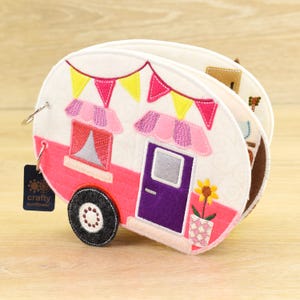 May include: A handmade, fabric camper-shaped book with a pink and white exterior. The camper features a purple door, a window with a red curtain, and a black wheel. A banner of pink and yellow flags decorates the top.