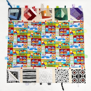 May include: A colorful baby activity quilt with various textures and patterns. The quilt features a central panel with a repeating pattern of airplanes, trucks, and the words "FLY", "DIG", "STOP", and "GO". The quilt has multiple flaps with different textures and patterns, including a patchwork design, a polka dot pattern, and a geometric pattern.