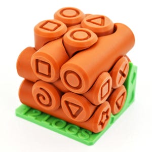 以下が含まれることがあります： オレンジ色の3Dプリントパズルキューブ。ゲームコントローラーのボタンのシンボルが特徴です。 キューブは「12 LOGS」の文字が入った緑色のベースの上にあります。 正方形、円、三角形、十字などの形状が含まれています。