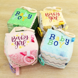 Può includere: Quattro cubi per bambini colorati con testo ricamato e anelli attaccati. I blocchi sono verdi, gialli, rosa e blu, con le parole "Baby Boy" e "Baby Girl". Ogni blocco ha un motivo e una consistenza diversi.