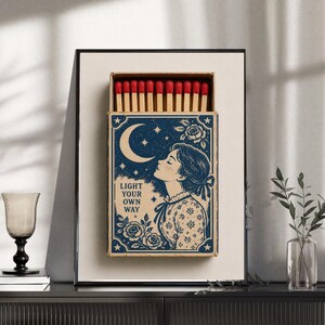 May include: A framed wall art piece featuring a matchbox design with a blue and cream illustration of a person looking at a crescent moon and stars. The text reads LIGHT YOUR OWN WAY. The matchbox is filled with wooden matches with red tips.