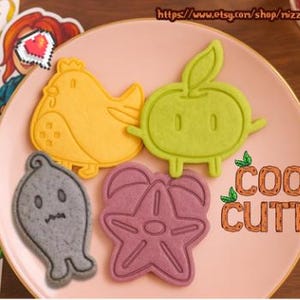 May include: A pink plate holds four colorful cookie cutters: a yellow chicken, a green apple, a gray ghost, and a purple starfish. The words "COOKIE CUTTERS" are written in orange. A calendar and sticker are in the background.