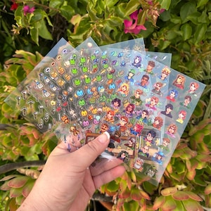 May include: Several sheets of clear stickers featuring colorful pixel art characters and objects. The stickers are held in front of a background of green foliage. The stickers include images of people, musical instruments, and other decorative elements.