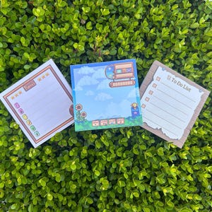 May include: Three square notepads with different designs. One has a To Do List, another has a pixelated computer and sky scene, and the third has a checklist with stars. All are on a green leafy background.