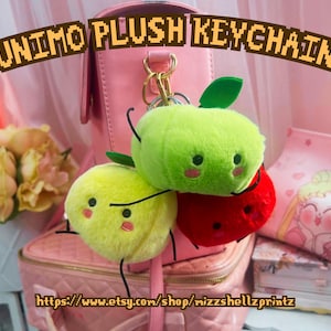 May include: Plush Junimo keychains in the shape of green, yellow, and red apples, each with black eyes and stems. The keychains are attached to a gold-coloured clasp. The background features a pink bag, flowers, and a pink pouch with a cartoon character.