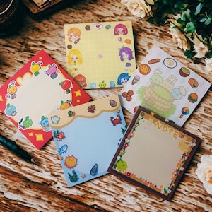 May include: A collection of colorful square sticky notes with various designs. One features cartoon characters, another has food illustrations, and others have decorative borders with cute graphics. The notes are arranged on a wooden surface with pens and flowers.