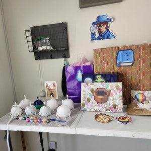 Pu&ograve; includere: Un tavolo con una variet&agrave; di regali avvolti in carta colorata. Ci sono anche diverse piccole figure di ceramica bianca con vernice e pennelli sul tavolo. I regali includono una scatola con un personaggio pixelato blu su di essa, una scatola con un personaggio pixelato marrone su di essa e una scatola con un personaggio pixelato arcobaleno su di essa. C'&egrave; anche un quadro incorniciato con il testo "L'avventura ti aspetta" su di esso.