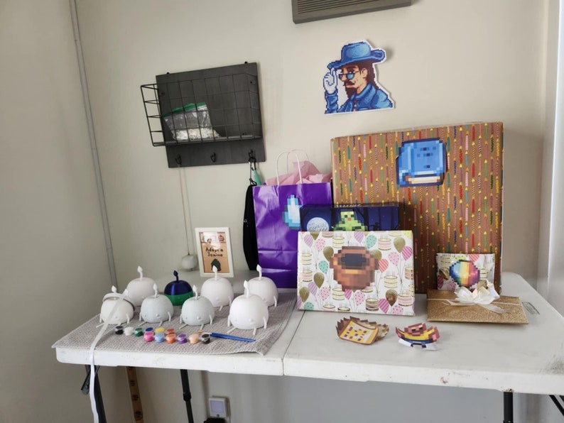Pu&ograve; includere: Un tavolo con una variet&agrave; di regali avvolti in carta colorata. Ci sono anche diverse piccole figure di ceramica bianca con vernice e pennelli sul tavolo. I regali includono una scatola con un personaggio pixelato blu su di essa, una scatola con un personaggio pixelato marrone su di essa e una scatola con un personaggio pixelato arcobaleno su di essa. C'&egrave; anche un quadro incorniciato con il testo "L'avventura ti aspetta" su di esso.