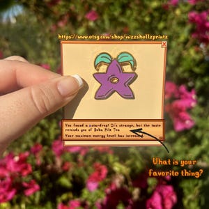 May include: A purple star-shaped enamel pin with teal accents, set on a tan background. The pin features text that reads, "You found a a stardrop!" and "What is your favorite thing?" The Etsy shop URL is visible.