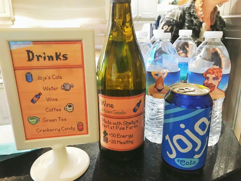 Pu&ograve; includere: Un cartello con la parola "Drinks" in lettere pixelate, che elenca Joja's Cola, Water, Wine, Coffee, Green Tea e Cranberry Candy. Una bottiglia di vino con un'etichetta che recita "Wine Artisan Goods Made with Shelly's feet at Pink Farm +50 Energy +20 Healthy". Una lattina di Joja Cola.