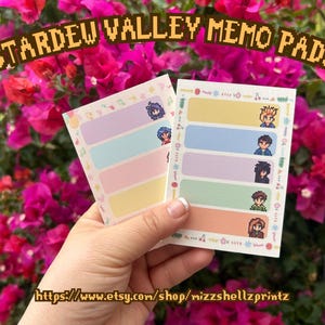 May include: Two memo pads with pastel colored sections and pixelated character art. The pads are labeled "Stardew Valley Memo Pads" in a pixelated font. The background features vibrant pink flowers. The Etsy shop URL is visible at the bottom.