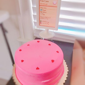 Pu&ograve; includere: Una torta rosa con caramelle a forma di cuore sulla cima. La torta &egrave; su un supporto per torte dorato. Un cartello accanto alla torta dice "Pink Cake" e elenca gli ingredienti e le informazioni nutrizionali. Il cartello dice "Store Bought", "Other Info: Sugar, Wheat, Egg", "There's little heart candies on top", "+250 Energy", "-112 Healthy".