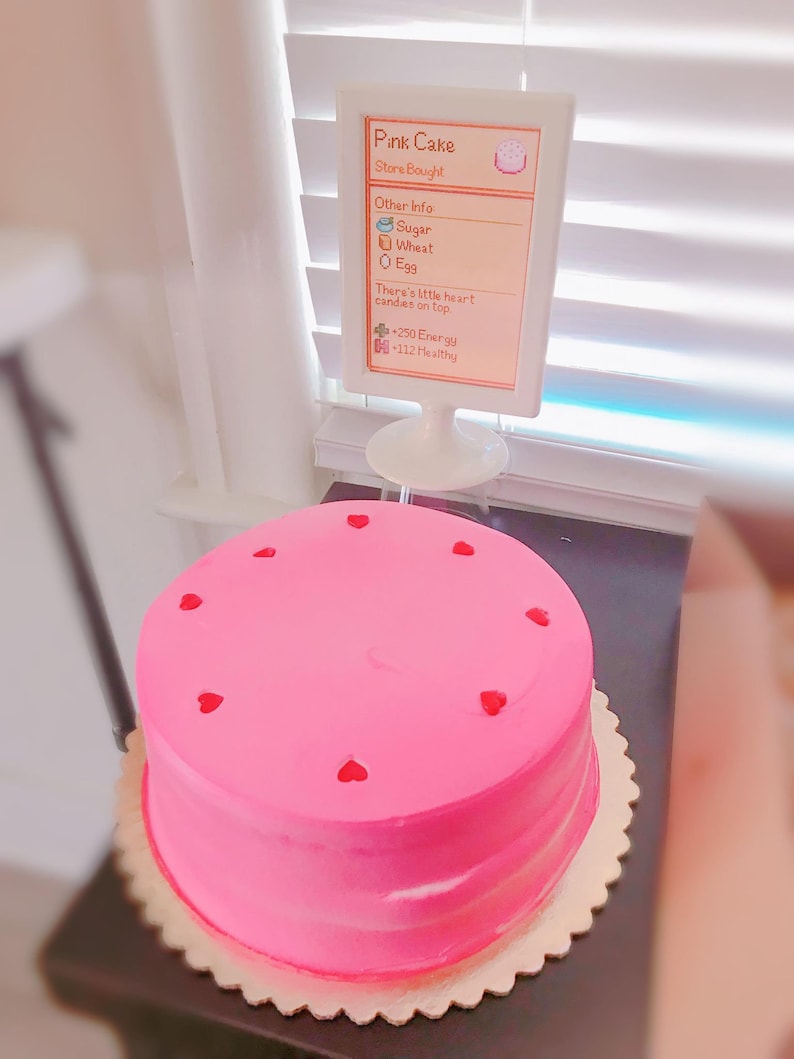 Pu&ograve; includere: Una torta rosa con caramelle a forma di cuore sulla cima. La torta &egrave; su un supporto per torte dorato. Un cartello accanto alla torta dice "Pink Cake" e elenca gli ingredienti e le informazioni nutrizionali. Il cartello dice "Store Bought", "Other Info: Sugar, Wheat, Egg", "There's little heart candies on top", "+250 Energy", "-112 Healthy".