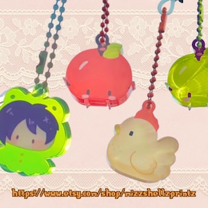 Puede incluir: Cuatro colgantes de acrílico de colores colgando de cadenas de bolas. Los colgantes incluyen un personaje verde con forma de rana, una manzana roja, un pollito amarillo y una cebolla verde. El fondo es de color rosa claro con un diseño de encaje. Texto abajo: https://www.etsy.com/shop/wizzshellzprintz