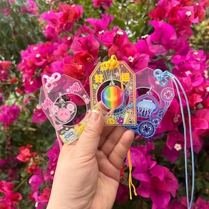 May include: Three clear acrylic charms with colourful designs are held in front of a vibrant pink flower bush. The charms feature a pig, a rainbow orb, and a jellyfish. Each charm has a different coloured string attached for hanging.