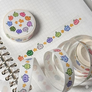 May include: A roll of decorative washi tape featuring a repeating pattern of colorful cartoon characters and star shapes. The tape is white with illustrations in shades of green, orange, pink, blue, and purple. The tape is unspooled, revealing the pattern.