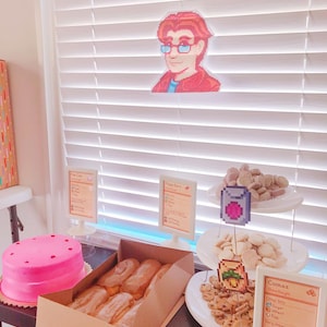 Pu&ograve; includere: Una torta rosa con glassa bianca e codette rosse si trova su un tavolo accanto a una scatola di pasticcini lunghi e marroni. Un supporto a tre livelli contiene vari biscotti, tra cui biscotti al cioccolato e biscotti allo zucchero. Il supporto &egrave; decorato con immagini pixelate di un gioiello rosa e una pianta di grano. Tre cartelli con grafica e testo pixelati sono sul tavolo. I cartelli indicano "Crosta di torta", "Barrette d'acero" e "Biscotti".