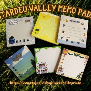 May include: A collection of Stardew Valley memo pads with various designs, including a star, cloud, and to-do list. The pads feature colorful illustrations and the text "Stardew Valley Memo Pads" and "Daily Plan". The pads are arranged on a bed of green grass.