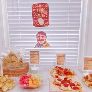 Pu&ograve; includere: Un tavolo con una variet&agrave; di cibi, tra cui patatine fritte, insalata di frutta, pizza e mini hamburger. Il tavolo &egrave; decorato con un cartello che dice "The Stardrop Saloon".