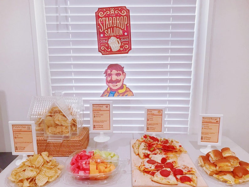 Pu&ograve; includere: Un tavolo con una variet&agrave; di cibi, tra cui patatine fritte, insalata di frutta, pizza e mini hamburger. Il tavolo &egrave; decorato con un cartello che dice "The Stardrop Saloon".