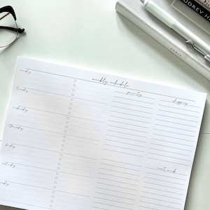 May include: A white weekly schedule notepad with sections for Monday through Sunday, priority, and shopping. The notepad has a black line border and a handwritten title "weekly schedule".