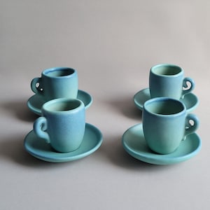 May include: Four small, turquoise ceramic espresso cups and saucers. Each cup has a rounded handle and sits on a matching saucer. The cups and saucers have a matte finish and a gradient of blue and turquoise colors.